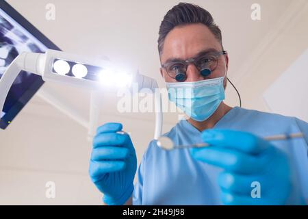 Dentiste mâle caucasien portant un masque facial tenant des outils dentaires à la clinique dentaire moderne Banque D'Images