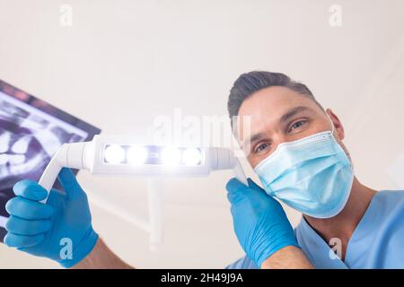 Dentiste mâle caucasien portant un masque facial tenant une lampe à la clinique dentaire moderne Banque D'Images