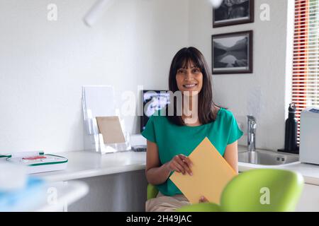Portrait d'une patiente biraciale souriante tenant des documents dans une clinique dentaire moderne Banque D'Images