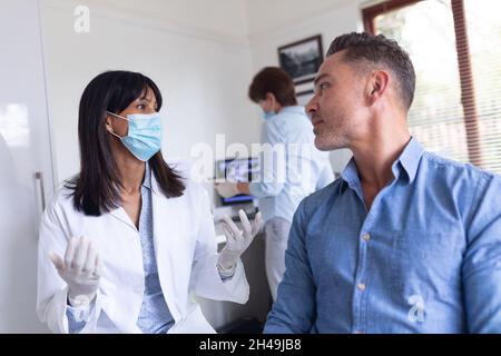 Dentiste biracial féminin portant un masque facial et parlant avec un patient masculin à la clinique dentaire moderne Banque D'Images