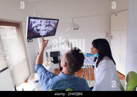 Dentiste biracial femelle portant un masque facial examinant les dents d'un patient mâle à la clinique dentaire moderne Banque D'Images