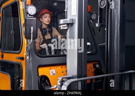 Travailleur féminin focalisé conduisant une chargeuse dans un entrepôt de conteneurs industriel, en toute confiance, vêtu d'un uniforme d'ingénieur et d'un casque orange rigide, conc Banque D'Images