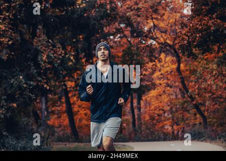 Un homme à la peau brune en bonne santé et en forme court dans les bois sur un sentier pavé dans une veste noire et un bonnet en automne et en automne, jogging et entraînement Banque D'Images