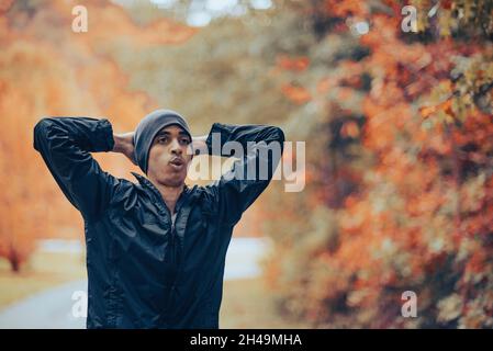 Beau homme de course mixte en bonnet gris s'étire, respire et se repose après un jogging ou un entraînement cardio dans le parc à l'automne.Respiration après l'entraînement Banque D'Images