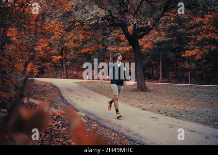 Un jeune homme à la peau brune court sur un sentier dans un parc entouré de feuilles d'automne.L'homme afro-américain se présente dans un parc à l'automne avec un bonnet noir Banque D'Images