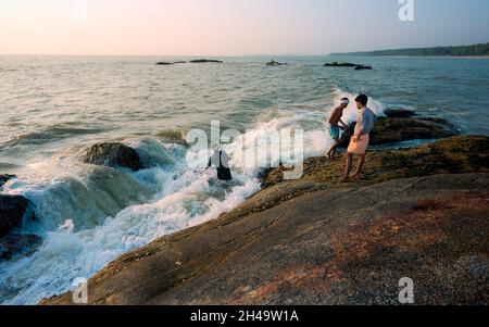 Des hommes locaux pêchent des coquillages provenant de grands rochers et entre les vagues sur la mer d'Arabie près de la côte au coucher du soleil près de Kannur, Kerala, Inde. Banque D'Images