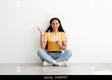 Offre exceptionnelle.Portrait d'une jeune femme arabe excitée avec un smartphone dans les mains assis sur le sol près du mur blanc, surprise Brunette Lady appréciant New Mobi Banque D'Images