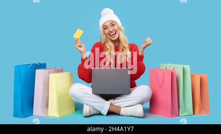 Wow, grosses ventes.Une jolie dame excitée dans un chapeau blanc faisant l'achat en ligne sur un pc, assis sur le sol avec des sacs d'achats, levant les mains vers le haut secouer les poings, tenir Banque D'Images