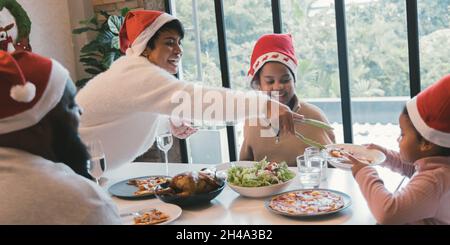 Belle jeune famille afro-américaine de mère, de père et de deux filles portant un chapeau de père noël, pour un dîner de noël avec pizza et dinde à la maison Banque D'Images