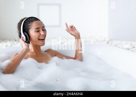 Belle jeune femme portant des écouteurs et chantant tout en étant couché dans un bain bouillant chaud à la maison, espace de copie.Belle femme millénaire à l'écoute de musique Banque D'Images