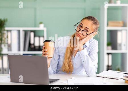 Portrait d'un jeune employé de bureau fatigué assis au travail avec un ordinateur portable tout en tenant une tasse de café et en regardant l'appareil photo, se sentant rouillé après le travail Banque D'Images