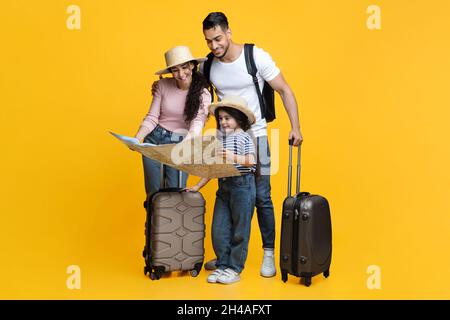 Bonne famille jeune avec petite fille regardant la carte, planification de voyage tout en se tenant debout avec des valises sur fond de studio jaune, parents arabes et Banque D'Images