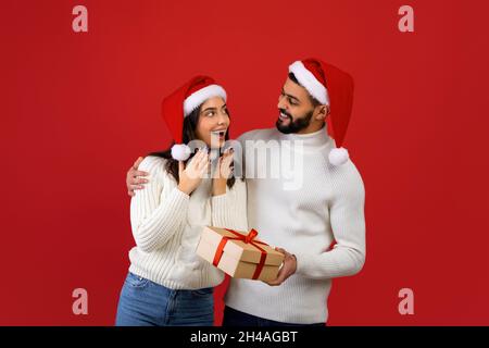 Cadeaux de Noël.Un mari arabe aimant surprend une jeune femme, donnant une boîte cadeau enveloppée et embrassant, portant des chapeaux de Père Noël tout en se tenant sur le dos rouge Banque D'Images