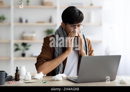 Photo d'un jeune homme d'affaires indien malade avec foulard autour du cou assis au bureau devant un ordinateur portable, regardant l'écran de portable, éternuant, s'est mis si Banque D'Images
