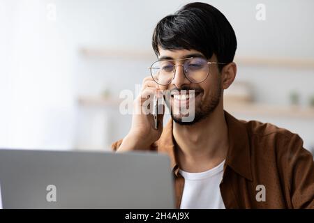 Gros plan portrait d'un beau gars indien avec des lunettes freelance travaillant à la maison, assis devant un ordinateur portable et ayant conversation téléphonique avec un clien Banque D'Images