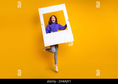 Vue sur toute la longueur du corps de fille gaie jumping attrayante tenant grand cadre de papier ayant amusant isolé sur fond jaune vif Banque D'Images