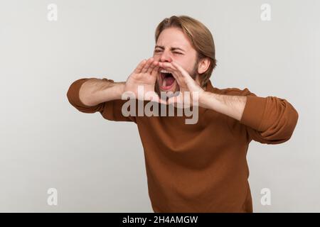 Portrait d'un homme furieux et séduisant avec une barbe portant un sweat-shirt, criant bruyamment dans la panique, tenant les mains près de sa bouche, faisant une annonce importante.Prise de vue en studio isolée sur fond gris. Banque D'Images