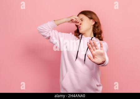 Portrait d'une jeune fille mécontentée dans la grimace à capuche dans le dégoût et la tenue de souffle, saisissant le nez et montrant l'arrêt pour éviter l'odeur pieuse, gaz fart.Studio d'intérieur isolé sur fond rose Banque D'Images