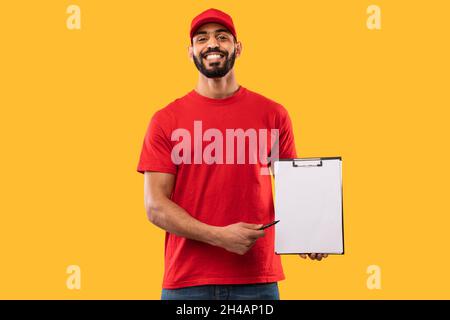 Souriant Arab Delivery Guy en Rouge uniforme montrant le presse-papiers à la caméra debout posant sur le fond jaune de studio.Publicité de service de messagerie, expédition Banque D'Images