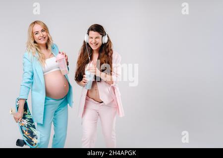 Deux filles enceintes en costume turquoise et rose avec des verres de jus, un skate et un casque se tiennent sur un fond gris. Banque D'Images