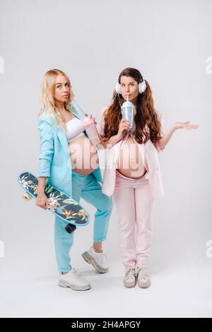 Deux filles enceintes en costume turquoise et rose avec des verres de jus, un skate et un casque se tiennent sur un fond gris. Banque D'Images