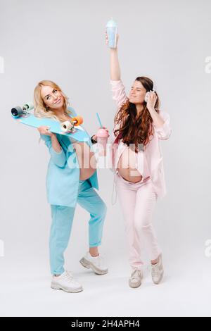 Deux filles enceintes en costume turquoise et rose avec des verres de jus, un skate et un casque se tiennent sur un fond gris. Banque D'Images