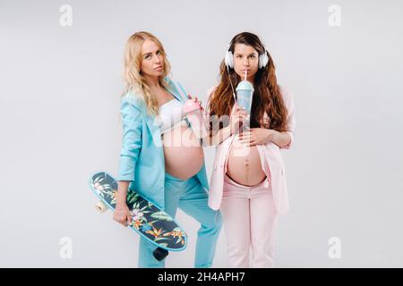 Deux filles enceintes en costume turquoise et rose avec des verres de jus, un skate et un casque se tiennent sur un fond gris. Banque D'Images