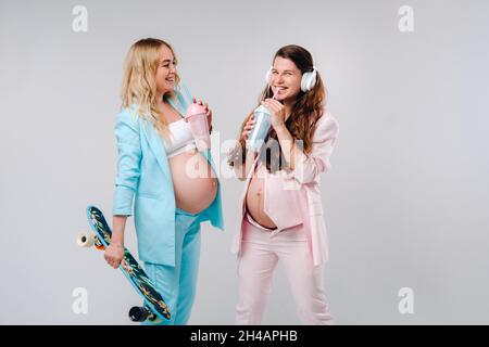 Deux filles enceintes en costume turquoise et rose avec des verres de jus, un skate et un casque se tiennent sur un fond gris. Banque D'Images