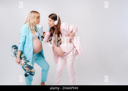 Deux filles enceintes en costume turquoise et rose avec des verres de jus, un skate et un casque se tiennent sur un fond gris. Banque D'Images