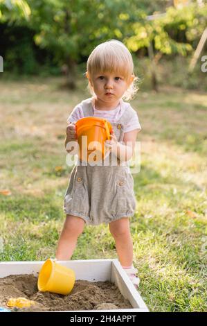 Une petite fille sérieuse dans une combinaison grise avec short avec un seau de jouet orange avec le sable dans ses mains joue dans le jardin.Il y a d'autres jouets Banque D'Images