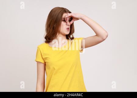 Troublée malheureuse adolescente attrayante fille avec des cheveux ondulés bruns saisissant le nez avec les doigts et étirant la main dehors sentiment dégoûté odeur désagréable.Prise de vue en studio isolée sur fond gris. Banque D'Images