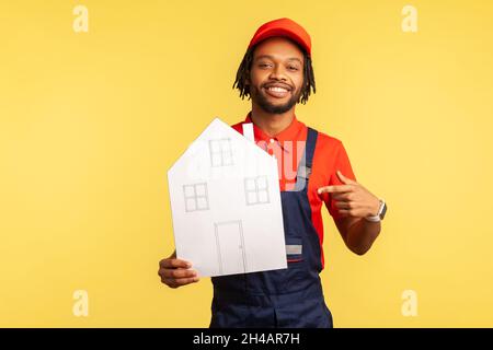 Homme à barbe souriant portant un T-shirt rouge et une combinaison bleue, pointant du doigt vers la maison de papier dans sa main, regardant l'appareil photo avec un sourire crasseux.Studio d'intérieur isolé sur fond jaune. Banque D'Images