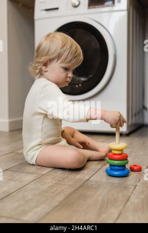 Un petit enfant est assis sur le sol dans la buanderie de la maison et joue avec un jouet multicolore pour enfants - un constructeur de pyramide Banque D'Images