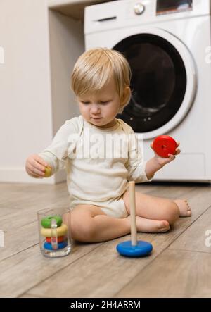 Un petit enfant est assis sur le sol de la maison et met des anneaux colorés d'une pyramide de jouets en bois pour enfants dans une tasse en verre.Style de vie Banque D'Images