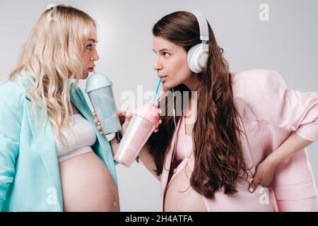 Deux filles enceintes en costume turquoise et rose avec des verres de jus, un skate et un casque se tiennent sur un fond gris. Banque D'Images