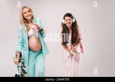 Deux filles enceintes en costume turquoise et rose avec des verres de jus, un skate et un casque se tiennent sur un fond gris. Banque D'Images