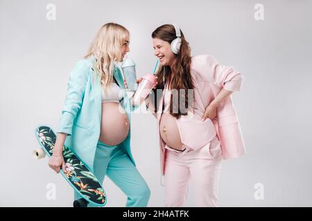 Deux filles enceintes en costume turquoise et rose avec des verres de jus, un skate et un casque se tiennent sur un fond gris. Banque D'Images