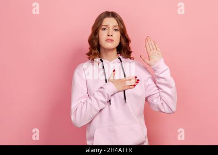 Portrait d'une jeune fille sincère et honnête à capuche tenant la main vers le haut et sur la poitrine, en juchant allégeance, en prêtant serment d'expression responsable.Studio d'intérieur isolé sur fond rose Banque D'Images