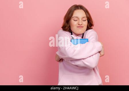 Portrait de la bonne brune adolescente fille à capuche embrassant cadeau d'anniversaire serré, souriant avec plaisir, regardant heureux rêveur, le meilleur présent jamais.Studio d'intérieur isolé sur fond rose Banque D'Images