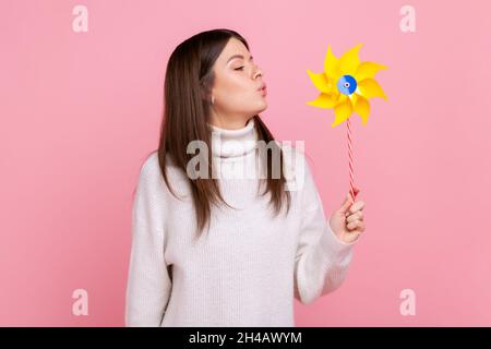 Portrait de femme brune sans souci soufflant à papier moulin à vent, jouant avec le jouet de pinwheel sur bâton, portant blanc style décontracté pull.Studio d'intérieur isolé sur fond rose. Banque D'Images