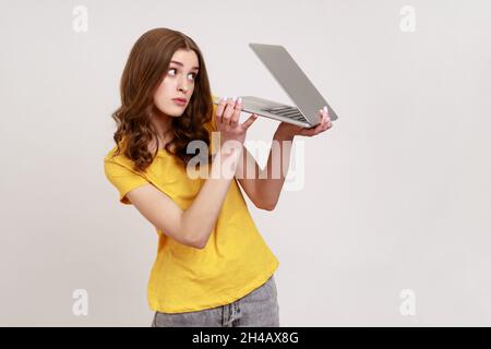Femme aux cheveux bouclés dans un T-shirt jaune de style urbain avec un ordinateur portable à demi-fermeture et un regard malicieux, espionnant et connaissant des informations secrètes.Prise de vue en studio isolée sur fond gris. Banque D'Images