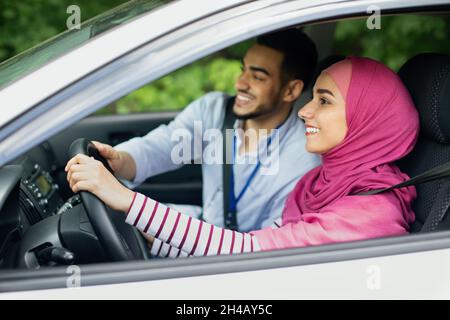Un mari arabe attentionné qui enseignait à sa femme musulmane dans le hijab à conduire sa voiture, un jeune homme du Moyen-Orient aidant le conjoint islamique à conduire un véhicule, tenant Steeri Banque D'Images
