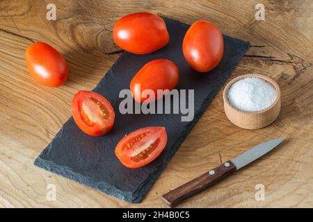 Beaucoup de tomates entières en forme de prune et une en tranches se trouvent dans une assiette et sur un tableau d'ardoise noire Banque D'Images