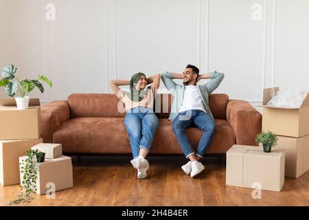 Un jeune mari et une femme arabes satisfaits se penchent sur le canapé, se reposent après avoir été déplacé, assis près des boîtes en carton et des plantes dans un nouvel appartement, copie s gratuite Banque D'Images