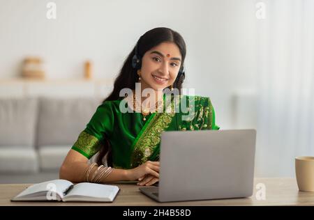 Portrait d'une femme indienne gaie en robe sari portant un casque, ayant une réunion en ligne ou une conférence Web sur ordinateur portable à la maison.Une étudiante prend p Banque D'Images
