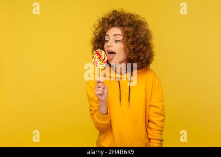 Femme attirante qui colle la langue à lécher, goûtant des sucreries rondes sucrées, savourant un dessert savoureux, portant un pull à capuche décontracté.Studio d'intérieur isolé sur fond jaune. Banque D'Images