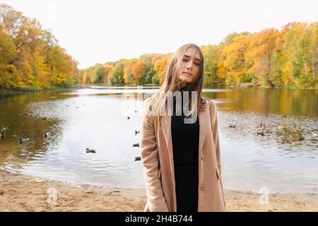 Jeune fille brunette appréciant l'automne au bord du lac en manteau de chameau dans le parc Banque D'Images