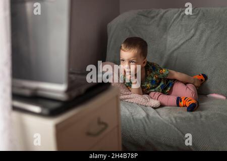 Petit garçon blond allongé sur un canapé et regardant des dessins animés sur les genoux Banque D'Images