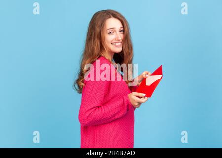 Portrait de la femme portant un pull-over rose, montrant une lettre romantique dans une enveloppe rouge, tenant le message d'amour et regardant l'appareil photo avec un sourire crasseux.Studio d'intérieur isolé sur fond bleu Banque D'Images