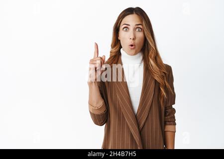 Portrait d'une femme excitée pdg, qui fait une idée, levant le doigt sur le geste d'eureka, montrant quelque chose sur le dessus, pointant vers le haut, se tenant sur un fond blanc Banque D'Images
